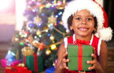 Excited African American child holding wrapped Christmas present gift box on Christmas day infront Christmas tree with Santa hat and smile.