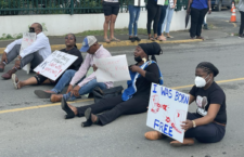 From left: Territorial At-Large Representative Neville 'Sheep' Smith, Sixth District Representative Alvera Maduro-Caines, social commentator Claude Skelton Cline, calypsonian Sistah Joyce, and Territorial At-Large Representative Sharie de Castro sit across a section of public road near Government House in Road Town, (Photo by Fitsroy Randall/BVI News)
