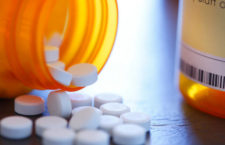 A close up of pills pouring out of a prescription medication bottle that rests on a table. The image is photogrphed using a very shallow depth of field.